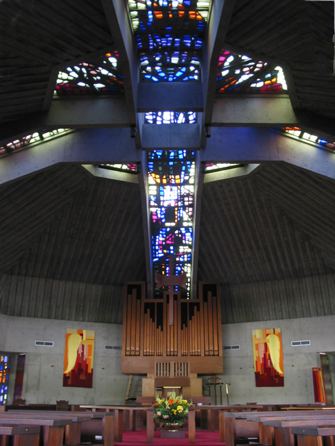 A massive stained glass and cement cross in the ceiling of a church full of wooden pews.
