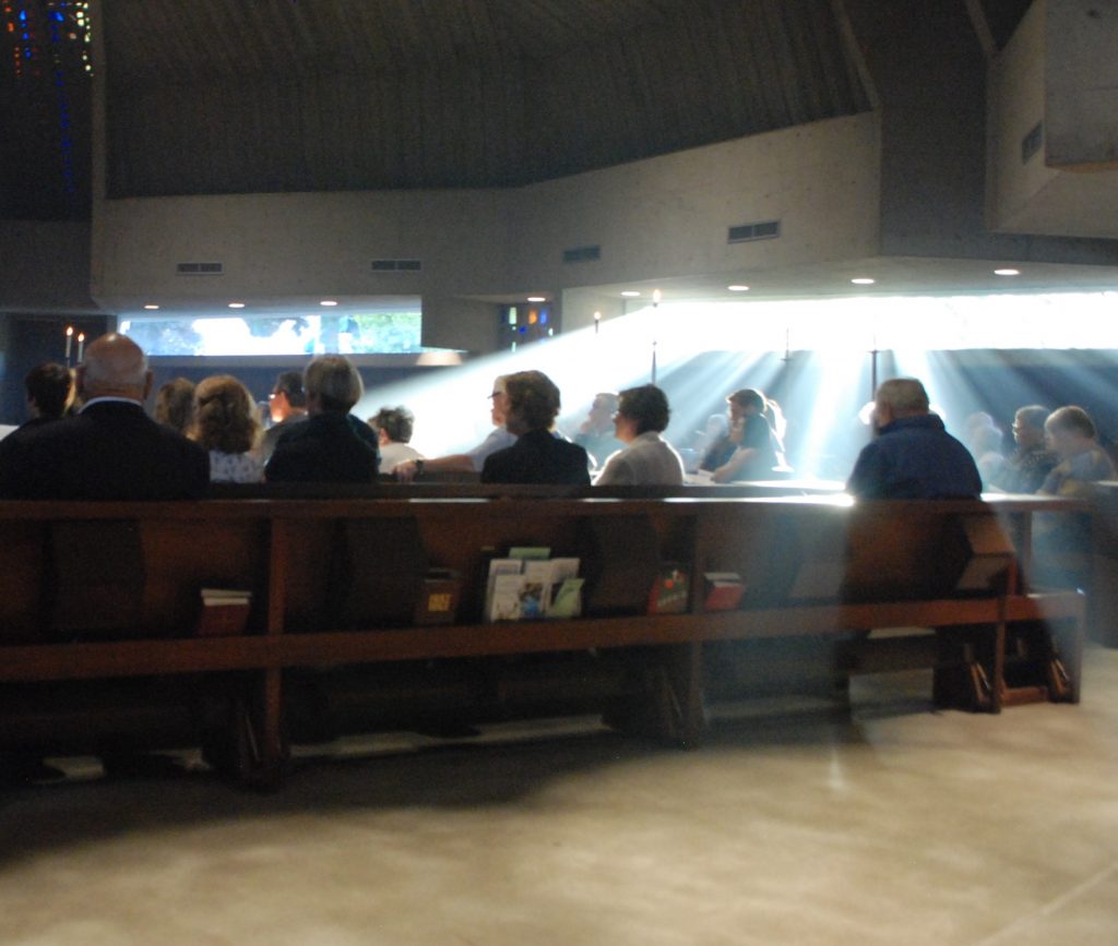 People sitting in pews with light streaming in over stained glass windows.