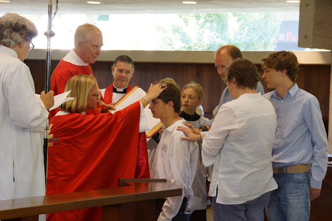 Bishop Mary Reaves kneeling while wearing red for Pentecost and putting her hand on a young person being confirmed in the church.