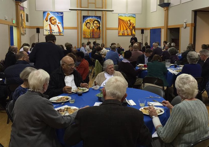 A group of adults eating around a table with banners depicting holy and working Latine people behind them.